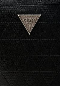 Fekete texturált kézitáska, melyen háromszög alakú, ezüst színű fém logó látható, rajta a "GUESS" felirattal és egy kérdőjellel, geometriai mintás háttér előtt.