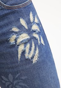 Siniset farkut, joissa on kulunut kuvio, rispaantuneet reunat ja vaaleaa kangasta paljastuu. Korostuksina on hienovarainen kukkakuosi.