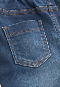 Tissu en denim avec une finition texturée, présente une petite poche arrière renforcée avec une couture orange contrastante et un design classique à cinq poches.