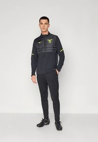 Fekete sportdzseki párnázott mellrésszel és függőleges csíkokkal, amelyeket neon sárga részletek kontrasztosítanak az ujjakon és a galléron. Fekete nadrág és cipők.