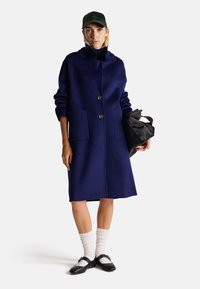 Cappotto di lana blu navy con colletto alto, due tasche frontali e bottoni marroni. Abbinato a calze bianche a coste e scarpe Mary Jane nere.
