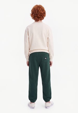 Enfant aux cheveux roux bouclés portant un sweat-shirt crème, un pantalon vert foncé avec un logo sur la poche arrière, et des baskets roses claires, debout, de dos.