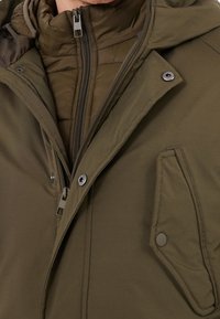 Chaqueta aislante de color verde oliva con cierre frontal de cremallera, cuello alto, botones a presión y bolsillo en el pecho, con un acabado de tejido texturizado.