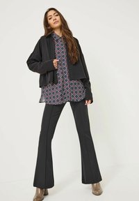 Schwarze kurze Jacke über einem gemusterten Hemd mit roten und blauen geometrischen Formen. Schwarze ausgestellte Hosen und beigefarbene Stiefeletten mit Absatz runden das Outfit ab.