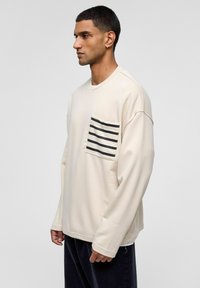Beige oversized sweatshirt met lange mouwen, voorzien van een zwart-wit gestreepte zak op de linkerborst en een gladde textuur.