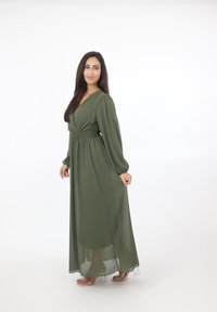 Vrouw met lang donker haar, gekleed in een lange olijfgroene jurk en transparante sandalen met hak, staand tegen een witte achtergrond.