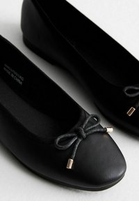 Ballerines noires en cuir avec un bout rond et une semelle flexible. Elles présentent un nœud décoratif noir avec des accents dorés à l'avant.