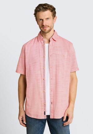 Mann mit kurzen lockigen Haaren, der ein pinkes kurzärmliges Hemd über einem weißen T-Shirt und einer blauen Jeans trägt, schaut nach vorne.