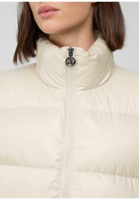 Žena vo výplnenej bunde krémovej farby s vysokým golierom zapnutá, ktorá zobrazuje čierny okrúhly detail na zips.