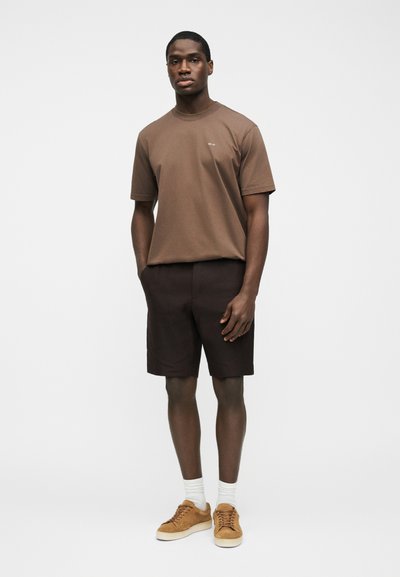 Άνδρας που στέκεται φορώντας καφέ κοντομάνικο μπλουζάκι, σκούρα καφέ σορτς, λευκές κάλτσες και ανοιχτό καφέ αθλητικά παπούτσια, μπροστά σε απλό φόντο.