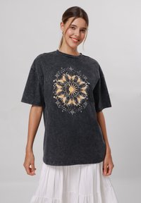 Oversized black t-shirt in washed cotton with a gold sun and butterfly graphic surrounded by stars and text on the front.