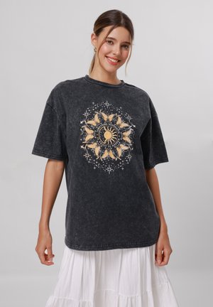T-shirt noir surdimensionné en coton lavé avec un motif doré de soleil et de papillon entouré d'étoiles et de texte sur le devant.