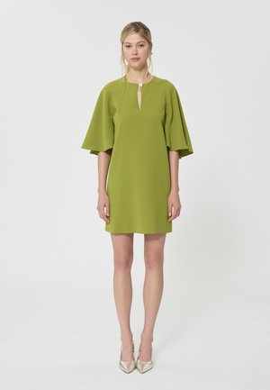 Femme debout portant une courte robe vert citron à larges manches jusqu'au coude et un col en trou de serrure avec un bouton décoratif.