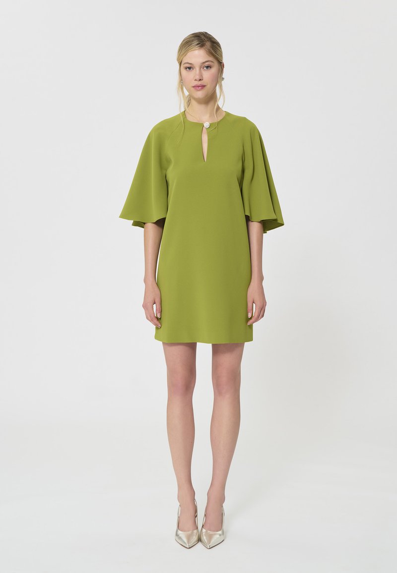 Femme debout portant une courte robe vert citron à larges manches jusqu'au coude et un col en trou de serrure avec un bouton décoratif.
