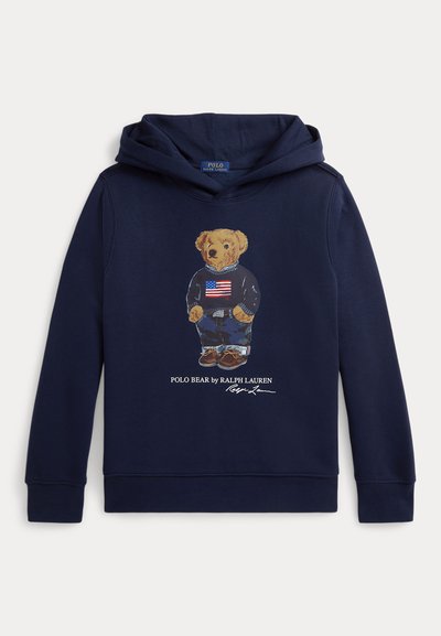 Laivastonsininen huppari, jossa karhun kuva, jonka päällä on neule, jossa on Amerikan lippu, farkut ja kengät, nimetty "Polo Bear by Ralph Lauren."
