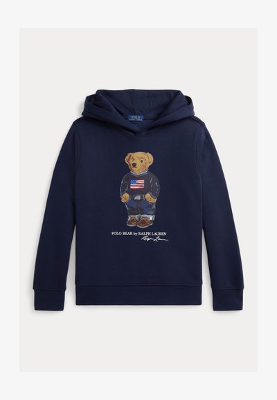 Felpa con cappuccio blu navy con illustrazione di un orso che indossa un maglione con una bandiera americana, jeans e scarpe, etichettata "Polo Bear by Ralph Lauren."