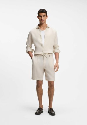 Mann trägt hellbeiges langärmeliges Hemd mit Knöpfen und hochgekrempelten Ärmeln, passende Shorts mit Kordelzug, weißes Unterhemd und schwarze Loafers.