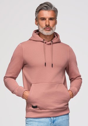 Homme aux cheveux gris et à la barbe portant un sweat à capuche rose poussiéreux avec poche avant et cordons, mains dans les poches, jean bleu clair.