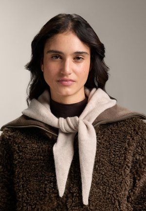 Barna, bolyhos dzseki magas gallérral, világos bézs sállal párosítva, ami csomózva van a nyaknál. Sima textúra és semleges árnyalatok.