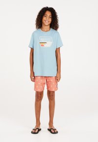 Lichtblauw T-shirt met "PROTEST" graphic, koraalkleurige zwembroek met witte palmbomenprint en zwarte slippers. Casual zomerse outfit.