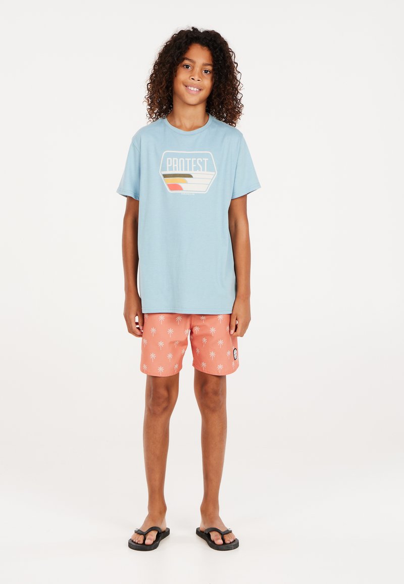 Lichtblauw T-shirt met "PROTEST" graphic, koraalkleurige zwembroek met witte palmbomenprint en zwarte slippers. Casual zomerse outfit.