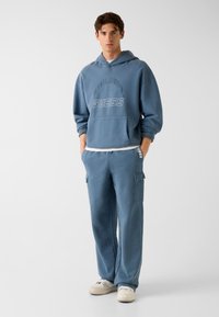 Niebieska bluza z kapturem z przednią kieszenią i logo "GUESS"; do kompletu jasnoniebieskie spodnie bojówki z kieszeniami po bokach; oba wykonane z miękkiego materiału.