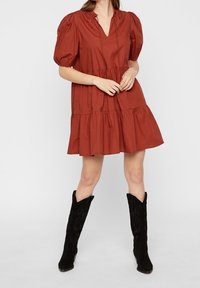 Vestido color óxido, a la altura de la rodilla, con mangas abullonadas y escote en V. Diseño en capas con frunce. Combinado con botas altas de ante negras.