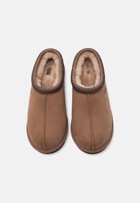 Chaussons en daim marron avec intérieur en peluche, bout arrondi et bordure tressée autour de l'ouverture. Présentent le logo UGG sur le côté.