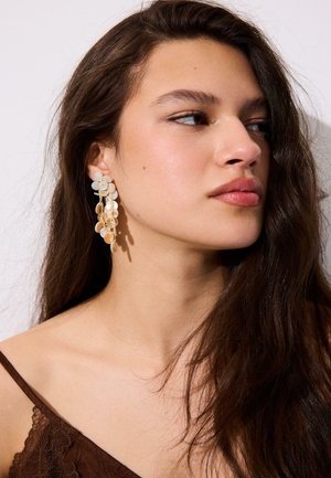 Mladá žena s dlhými tmavými vlasmi, ktorá nosí visiace kvetinové náušnice a hnedý top s čipkovou lemovkou, pozerá sa doprava na jednofarebnom pozadí.