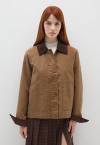 Braune Baumwolljacke mit einem Cordkragen, ausgestattet mit Druckknopfverschlüssen, zwei vorderen Taschen und weiten Manschetten. Getragen über einem karierten Rock.