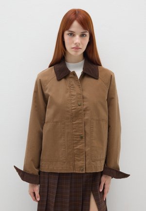 Braune Baumwolljacke mit einem Cordkragen, ausgestattet mit Druckknopfverschlüssen, zwei vorderen Taschen und weiten Manschetten. Getragen über einem karierten Rock.