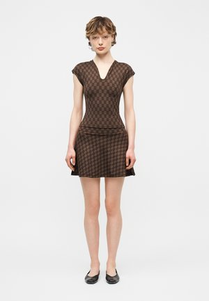Νεαρό άτομο που φοράει εφαρμοστό καφέ φόρεμα με σχέδιο, V-λαιμόκοψη και κοντά μανίκια, συνδυασμένο με μαύρα φλατ παπούτσια, στέκεται μπροστά σε απλό φόντο.