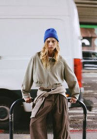 Oliven grøn cropped jakke med elastisk talje, kombineret med løse brune bukser. Modellen bærer en blå strikket hue og holder en sort stang.