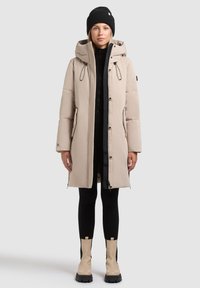 Manteau en duvet beige avec capuche, fermeture éclair et boutons-pression, accents à cordon de serrage, poches latérales, associé à des leggings noirs et des bottines beiges.