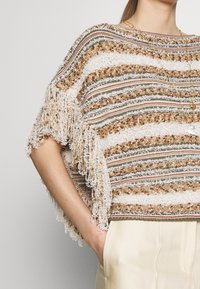 Strick-Poncho in Beige- und Erdtönen mit gestreiften Mustern, ausgestattet mit Fransen entlang des Saums und kurzen Ärmeln.