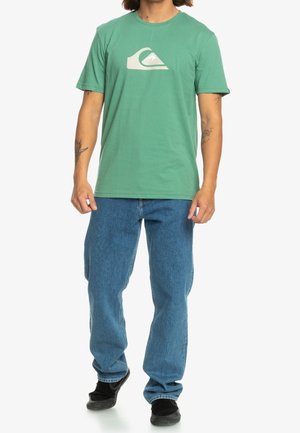 Hombre con camiseta verde de Quiksilver, jeans azules y zapatos negros, de pie con los brazos relajados a los lados.