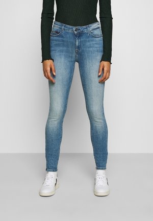 Modré džíny s úzkým střihem nošené s bílými teniskami a černým topem s dlouhým rukávem, zobrazené od poloviny trupu k nohám na jednobarevném pozadí.