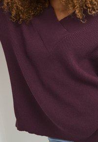 Lila rätstickad tröja med V-halsdesign, utrustad med texturerat tyg och en avslappnad passform. Nederkanten är lätt vågig.