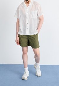 Muž v bílé krátké košili s tenkými pruhy, olivově zelených šortkách, bílých ponožkách a bílých teniskách na modré podlaze.