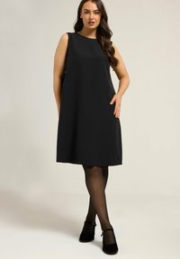 Vestido negro sin mangas en tejido suave, silueta en A, a la altura de la rodilla, con escote redondo y bolsillos laterales. Medias negras y zapatos.