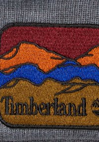 Parche bordado que presenta montañas en rojo, naranja, azul y marrón sobre un fondo gris, con "Timberland" en negro debajo.