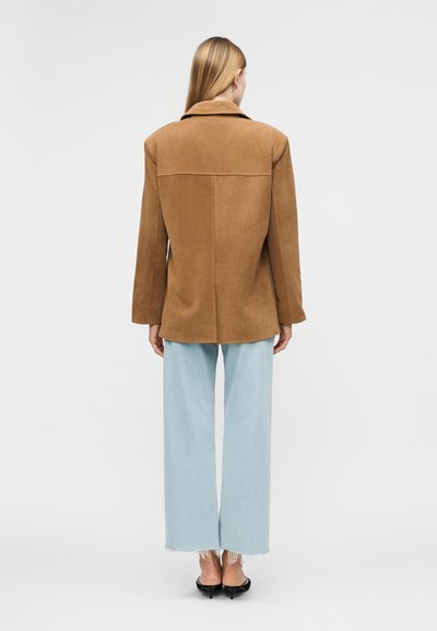 Γυναίκα στέκεται με την πλάτη στην κάμερα, φορώντας ένα καφέ δερμάτινο μπουφάν, φαρδιά τζιν σε ανοιχτό μπλε χρώμα και μαύρες γόβες με ανοιχτή φτέρνα πάνω σε λευκό φόντο.