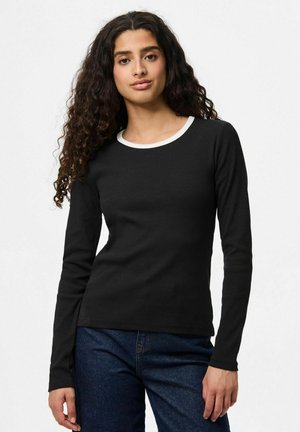 Vrouw met lang krullend haar draagt een zwarte lange mouw top met een witte rand bij de kraag en een blauwe jeans, staand tegen een effen lichte achtergrond.