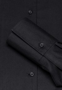 Camicia nera con bottoni sul davanti, tessuto morbido e polsini piegati con bottone abbinato, che mette in evidenza un design e una texture curati.