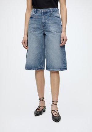 Femme portant un short en denim bleu clair à jambes larges et des ballerines pointues noires à lacets avec des détails cloutés.