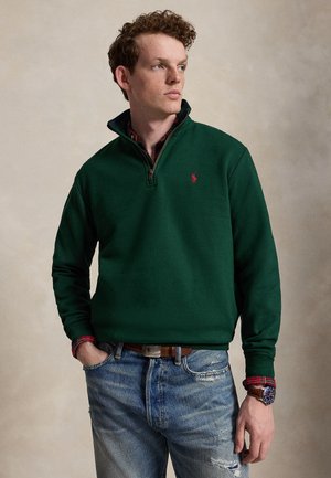 Jeune homme aux cheveux bouclés portant un sweat-shirt vert foncé à fermeture éclair quart de zip sur une chemise rouge à carreaux et un jean bleu, debout les mains dans les poches.