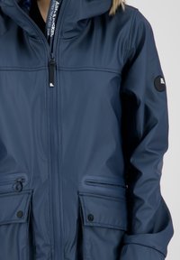 Wodoodporna granatowa kurtka z zamkiem z przodu, regulowanym kapturem, dwiema dużymi kieszeniami z przodu oraz okrągłym logo na lewym rękawie. Gładka faktura.