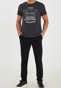 Svart t-shirt med vintage grafisk tryck i grått och vitt; ihop med svarta joggerbyxor; modell bär svarta sneakers.