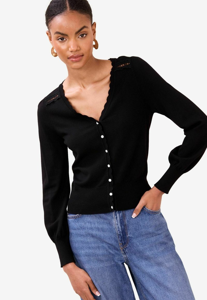 Cardigan noir avec col en V, bords festonnés, détails en dentelle sur les épaules et fermeture par boutons. Manches longues et taille cintrée.