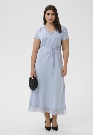 Vrouw met een lichtblauwe midijurk met kanten rand, taille met riem, een zwarte geweven clutch vasthoudend en zwarte puntige hakken dragend.